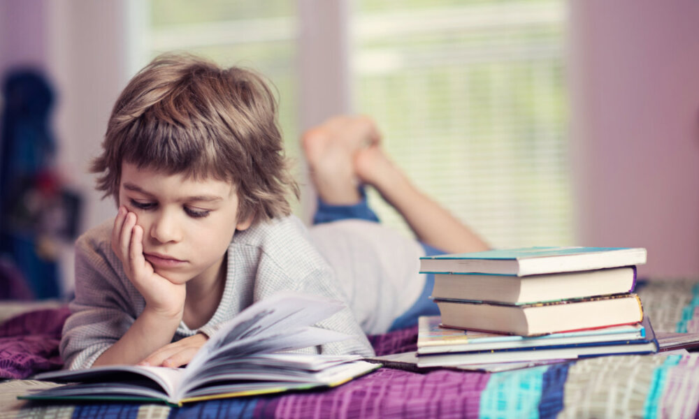 Cute little boy lying on bed reading next to stack of books