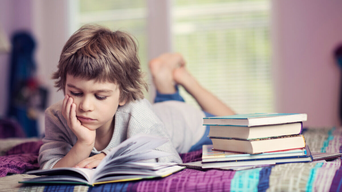 Cute little boy lying on bed reading next to stack of books