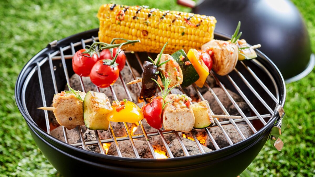 Vegetables and tofu kebab grilling on grid grille with fresh corn, viewed in close-up against green lawn grass in background