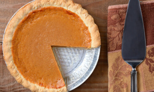 High angle shot of a pumpkin pie that has been cut into. Shot on a rustic wood table with a Autumn themed napkin and server by its side.