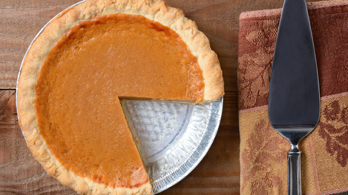 High angle shot of a pumpkin pie that has been cut into. Shot on a rustic wood table with a Autumn themed napkin and server by its side.