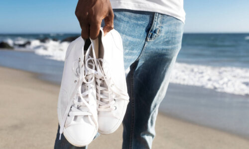 man holds pair of white sneakers on the beach